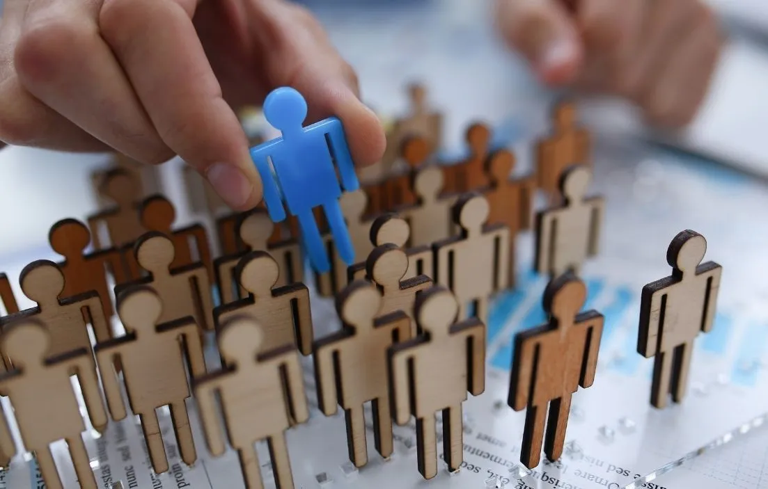 The hand of a person picking out a small blue person shaped cut out, from a small crowd of wooden people cut outs. Off a desk of paperwork.
