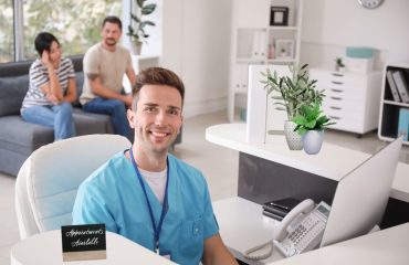Doctor sitting at the front desk of his medical office desk smiling at the camera, there are patients in the background and the office is modern.