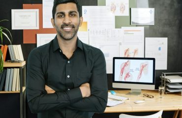 Male medical specialist in a black button up shirt leaning on his desk where you can see medical images like joints in the body on a pc and wall behind him.