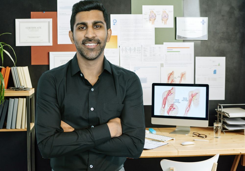 Male medical specialist in a black button up shirt leaning on his desk where you can see medical images like joints in the body on a pc and wall behind him.