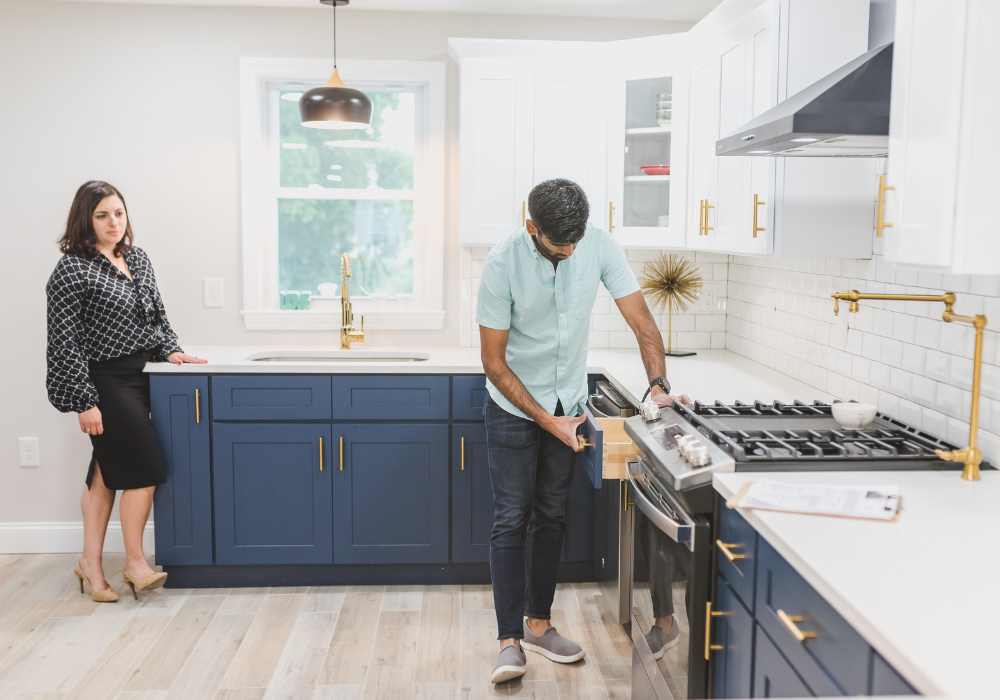 A man looking inside the kitchen drawers of a home he is looking to buy, with the female real estate agent looking on behind him.