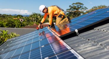 Man in a hard hat, orange workwear and harness, installing solar panels to a house roof in Brisbane