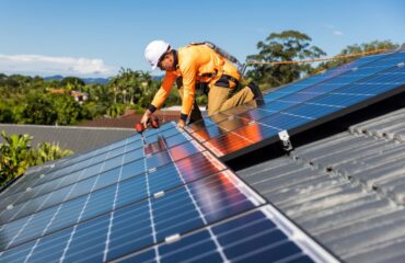 Man in a hard hat, orange workwear and harness, installing solar panels to a house roof in Brisbane