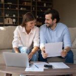 Man and women sit on a beige couch leaning over a coffee table and do their paperwork for their taxes.