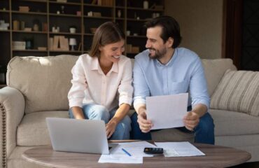 Man and women sit on a beige couch leaning over a coffee table and do their paperwork for their taxes.