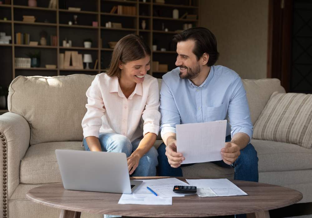 Man and women sit on a beige couch leaning over a coffee table and do their paperwork for their taxes.