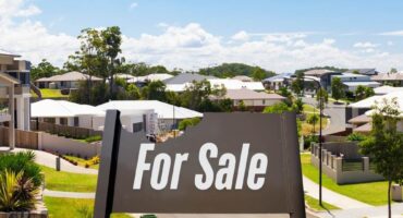 A brown for sale sign in front of a street of houses with a bite taken out if it.