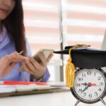 student at desk working out loan debt with a clock and graduation cap on the desk
