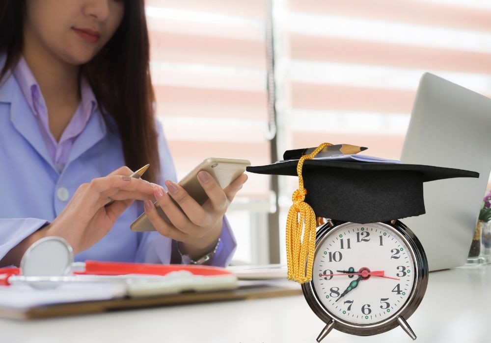 student at desk working out loan debt with a clock and graduation cap on the desk